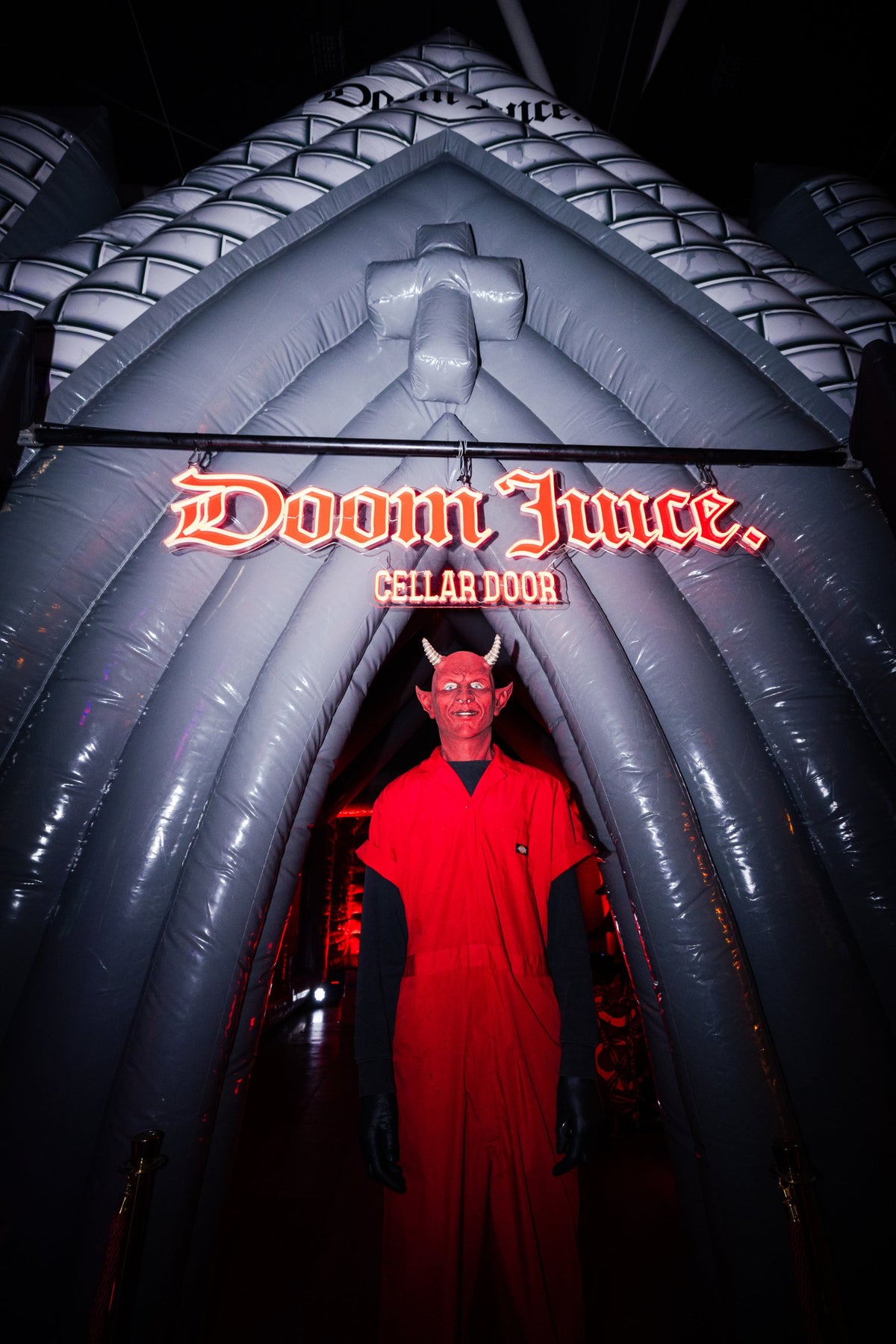 The Doom Juice inflatable church at night, with a red neon Doom Juice Cellar Door sign over the entrance and a devil standing in the doorway